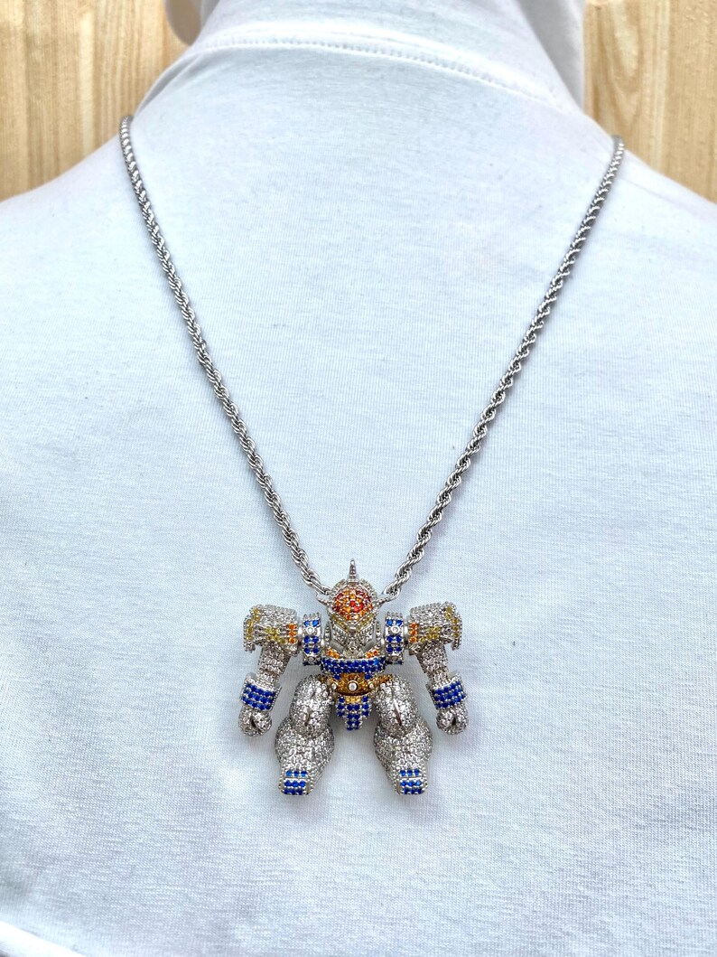 May include: Silver-toned robot pendant necklace. The robot pendant is encrusted with clear, blue, yellow, and orange gemstones. The necklace chain is a silver rope style.