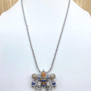 May include: Silver-toned rope chain necklace with a robot pendant. The robot pendant is encrusted with colorful gemstones in blue, yellow, orange, and clear. The robot has a detailed design with articulated limbs and a head.