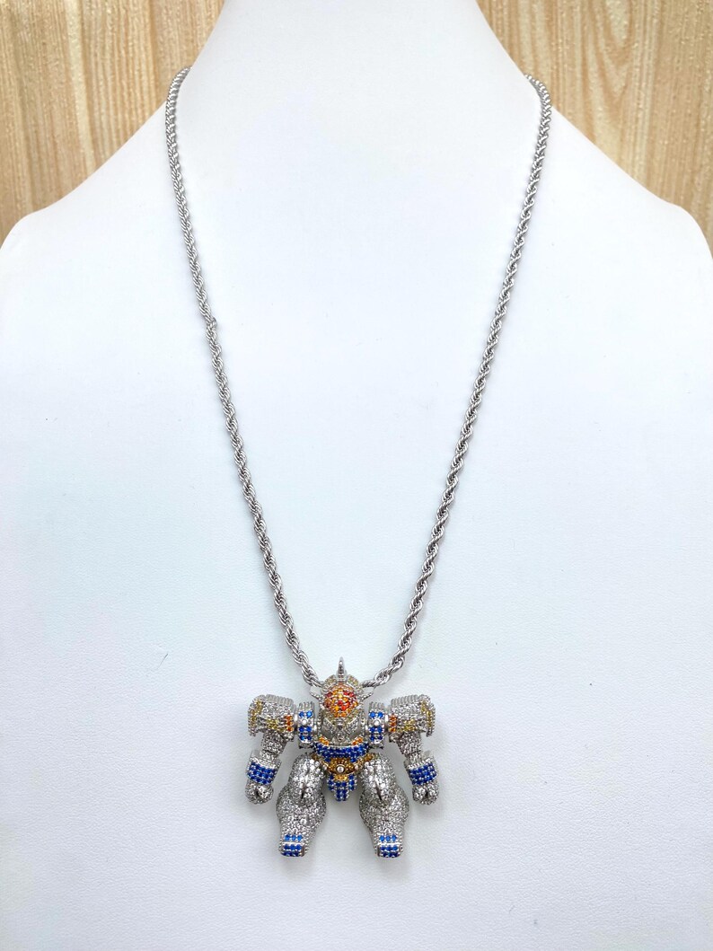 May include: Silver-toned rope chain necklace with a robot pendant. The robot pendant is encrusted with colorful gemstones in blue, yellow, orange, and clear. The robot has a detailed design with articulated limbs and a head.