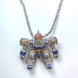 May include: Silver-toned robot pendant necklace with a rope chain. The robot is encrusted with clear, blue, yellow, and orange gemstones. The robot's head is orange and red, and the arms and legs are detailed with blue gems.
