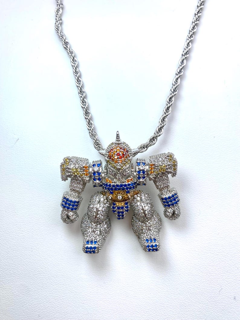 May include: Silver-toned robot pendant necklace with a rope chain. The robot is encrusted with clear, blue, yellow, and orange gemstones. The robot's head is orange and red, and the arms and legs are detailed with blue gems.
