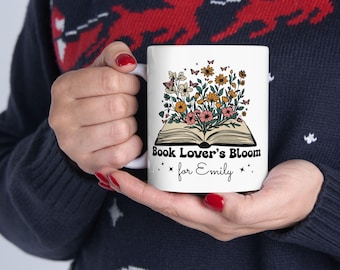 Può includere: Tazza in ceramica bianca con la scritta "Book Lover's Bloom" e un design floreale e a farfalle. La tazza è personalizzata con il nome "Emily". Il design include fiori nei toni del rosa, giallo e arancione.