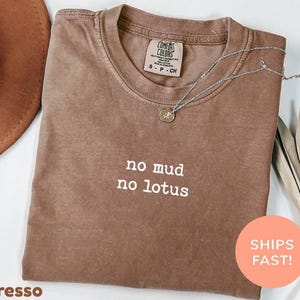 以下が含まれることがあります： エスプレッソ色のComfort Colors Tシャツ。白で「no mud no lotus」の文字。シルバーのネックレスがシャツの上に。茶色の帽子と乾燥した葉が背景にあります。