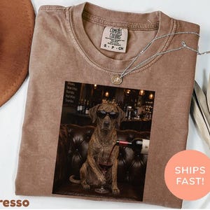 Puede incluir: Una camiseta marrón de cuello redondo con un estampado gráfico de un perro atigrado con gafas de sol negras, sentado en un sofá de cuero capitoné mientras sirve vino tinto en una copa. La camiseta tiene una etiqueta Comfort Colors en el cuello.