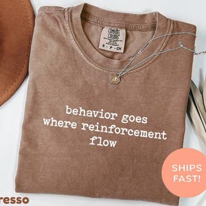 May include: Espresso-colored t-shirt with the text "behavior goes where reinforcement flow" in white. A silver necklace with a pendant rests on the shirt. The shirt has a Comfort Colors tag.