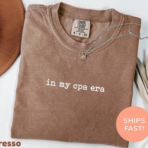 Könnte beinhalten: Espressofarbenes T-Shirt mit dem weißen Aufdruck "in my cpa era". Das Shirt hat einen Rundhalsausschnitt und ein kleines Etikett am oberen Rücken. Eine silberne Halskette mit Anhänger ist über dem Shirt drapiert. Ein runder Aufkleber mit den Worten "SHIPS FAST!" befindet sich unten rechts.