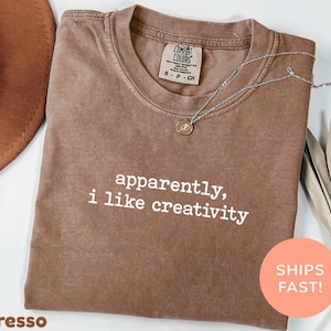 Puede incluir: Camiseta color espresso con el texto blanco "apparently, i like creativity". La camiseta tiene cuello redondo y mangas cortas. Un collar plateado con un colgante está drapeado sobre la camiseta. Un sombrero marrón y hojas secas están en el fondo.