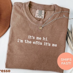 May include: Espresso-coloured t-shirt with the text "it's me hi i'm the efda it's me" in white. The shirt has a crew neck and short sleeves. A silver necklace with a small pendant is draped across the shirt.