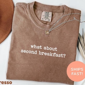 Op de afbeelding: Espresso-kleurig T-shirt met de witte tekst "what about second breakfast?". Het shirt heeft een ronde hals en een klein label op de bovenrug. Een zilveren ketting met een hanger is over het shirt gedrapeerd. Een ronde oranje sticker zegt "SHIPS FAST!"