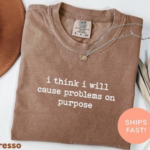 May include: Espresso-colored t-shirt with the phrase "i think i will cause problems on purpose" in white. A silver necklace with a pendant rests on the shirt. The shirt has a Comfort Colors tag.