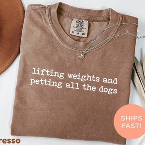 Puede incluir: Camiseta color espresso con el texto blanco "lifting weights and petting all the dogs". Un collar plateado con colgante descansa sobre la camiseta. La camiseta tiene cuello redondo y una etiqueta "Comfort Colors".