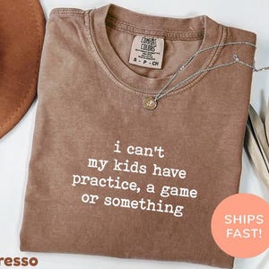 May include: Espresso-colored t-shirt with the text "i can't my kids have practice, a game or something" in white. A silver necklace with a pendant is draped across the shirt. The shirt has a Comfort Colors tag.