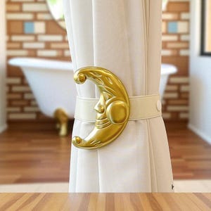 May include: A gold-colored crescent moon curtain tieback with a face design, holding a cream-colored curtain. The tieback is attached to the curtain with a cream-colored elastic band. The background shows a bathroom with a white bathtub.