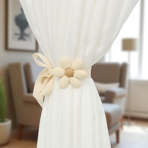 May include: Cream-colored curtain tieback featuring a daisy-shaped accent with a light brown center. The tieback is made of a soft, textured material and is tied with a matching ribbon. The curtains are white and sheer, adding a touch of elegance to the room.