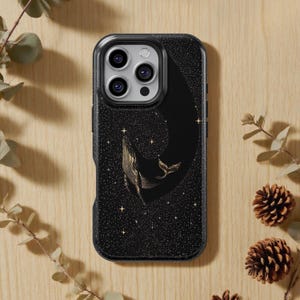 May include: Black phone case featuring a gold whale design against a starry night sky. The whale is depicted with intricate line art, and the case has a textured, glittery appearance. The case is shown on a light wood surface with sprigs and pinecones.