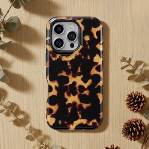 Puede incluir: Funda de teléfono inteligente con un diseño de carey en tonos marrón y amarillo. La funda tiene un borde negro y un recorte para la cámara. Descansa sobre una superficie de madera marrón claro, acompañada de ramitas verdes y piñas.