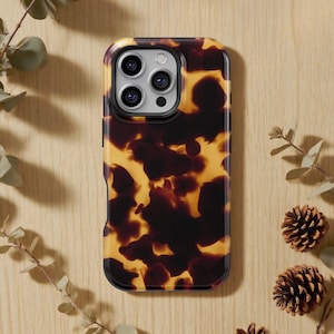 May include: A tortoiseshell-patterned phone case with a black border. The case has a glossy finish and is designed for a smartphone with multiple camera lenses. The background includes sprigs of greenery and pinecones.
