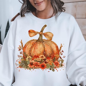 May include: White sweatshirt featuring a watercolor design of a large orange pumpkin with a bow and floral accents. The pumpkin has a glittery texture and is surrounded by autumn-colored flowers and foliage, perfect for fall.