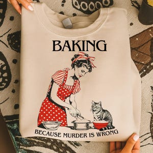 Può includere: Felpa beige con un'illustrazione retrò di una donna che cucina con un gatto. Il design include il testo "BAKING BECAUSE MURDER IS WRONG". La donna indossa un grembiule a pois rossi. Design vintage a tema cucina.