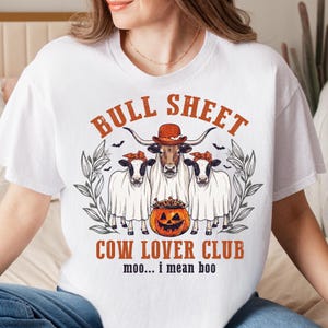 Può includere: Una t-shirt bianca con una grafica di tre mucche vestite da fantasmi, con la mucca centrale che indossa un cappello marrone. Sotto c'è una zucca di Halloween piena di caramelle. Il testo recita BULL SHEET COW LOVER CLUB e moo... i mean boo.