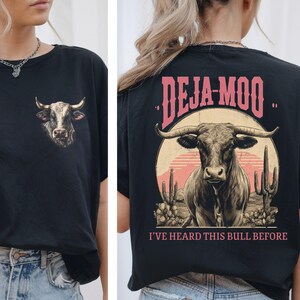 May include: Black t-shirt featuring a bull graphic on the front and back. The front has a bull's head, while the back displays a bull with the text "DEJA-MOO" and "I'VE HEARD THIS BULL BEFORE". The design includes a desert sunset and cacti.