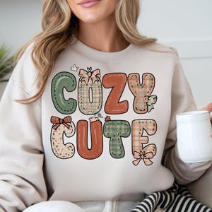 May include: A beige sweatshirt with the words "COZY CUTE" in a patchwork design. The letters are in green, orange, and tan with bow accents. The person is holding a white mug.
