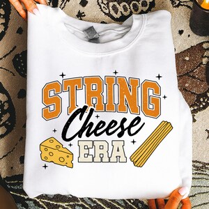 Könnte beinhalten: Weißes Sweatshirt mit dem Text "STRING Cheese ERA" in Orange und Schwarz. Das Design umfasst ein Käseeck und ein Bündel Käsefäden. Ein lustiges, essensbezogenes Grafikdesign für Freizeitkleidung.