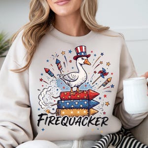 May include: Beige sweatshirt with a patriotic design. Features a white goose wearing a red, white, and blue top hat, standing on a stack of red, white, and blue rockets. The word "FIREQUACKER" is printed in black script below the rockets.