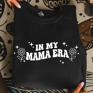 May include: Black sweatshirt with the text "IN MY MAMA ERA" in white, with flower and star graphics. The sweatshirt is folded and held by hands. The design has a retro, groovy aesthetic.
