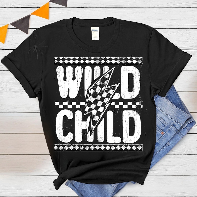 Puede incluir: Camiseta negra con la frase "WILD CHILD" en letras blancas. Un dise&ntilde;o de bandera a cuadros est&aacute; incorporado en un rayo. La camiseta se muestra con vaqueros azules sobre una superficie de madera blanca.