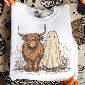 May include: White sweatshirt featuring a brown Highland cow and a beige ghost illustration. The cow has large horns and shaggy fur, while the ghost has a simple, smiling face. The design is set against a backdrop of tall grass.