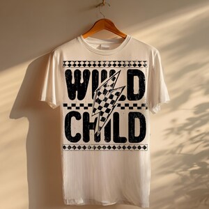 Puede incluir: Una camiseta blanca con las palabras "WILD CHILD" en negrita, negro, con un efecto desgastado. Un rayo a cuadros est&aacute; centrado entre las palabras. La camiseta est&aacute; colgada de una percha de madera.