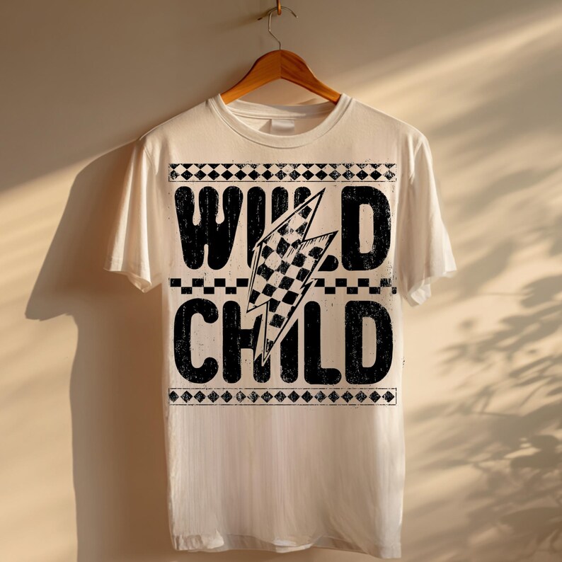 Puede incluir: Una camiseta blanca con las palabras "WILD CHILD" en negrita, negro, con un efecto desgastado. Un rayo a cuadros est&aacute; centrado entre las palabras. La camiseta est&aacute; colgada de una percha de madera.