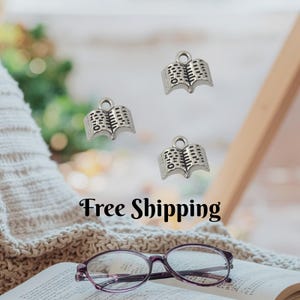 May include: Silver-colored open book charms with detailed page and spine designs. The charms are suspended in the air, with a pair of purple-framed glasses resting on an open book. The background is blurred, suggesting a cozy reading environment.