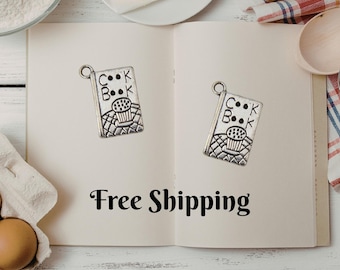 May include: Two silver-colored cookbook charms with a cupcake design are displayed on an open book. The book is open on a white wooden surface, with ingredients like flour, butter, and eggs in the background.