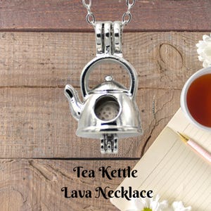May include: A silver-colored tea kettle pendant necklace. The pendant is shaped like a miniature teapot with a spout, handle, and a small opening. The necklace is on a silver chain. The text "Tea Kettle Lava Necklace" is below the pendant.