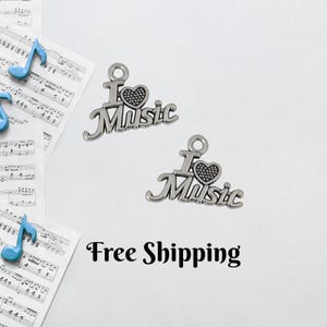 May include: Two silver-toned charms with the words "I Love Music" and a heart design. The charms are on a white surface with musical notes and the text "Free Shipping". The charms are likely for jewelry making or crafting.