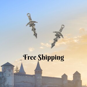May include: Two silver-colored dagger charms with ornate designs are suspended against a sky background. The daggers have detailed hilts and blades. The image also features a castle at the bottom.
