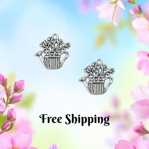 May include: Two silver-colored watering can charms, each with a plant design. The charms are detailed with vertical lines and heart-shaped accents. The background is a soft gradient of blue and pink with floral elements.