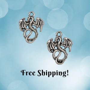 May include: Two silver-colored dragon charms with outstretched wings and curled tails. Each charm has a loop at the top for attaching to jewelry or other crafts. The background is a soft blue with blurred white circles.