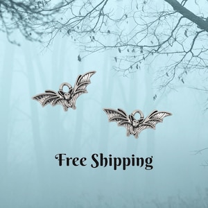 May include: Two silver-colored bat charms with detailed wing designs. Each bat has a small loop at the top for hanging. The image includes the text "Free Shipping" in a stylized font, set against a light blue background with tree branches.