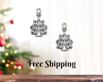 May include: Two silver-colored Christmas tree charms with intricate, openwork designs. Each charm has a loop at the top for hanging. The background is a blurred image of a Christmas tree and a wooden surface.