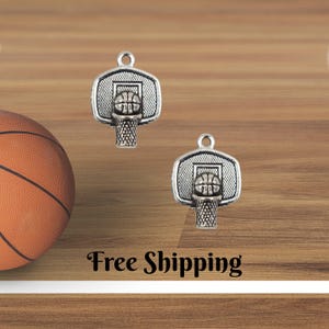 May include: Two silver-colored basketball hoop charms with a basketball and net design. A basketball is visible on the left. The text "Free Shipping" is at the bottom. The charms are on a wooden surface.