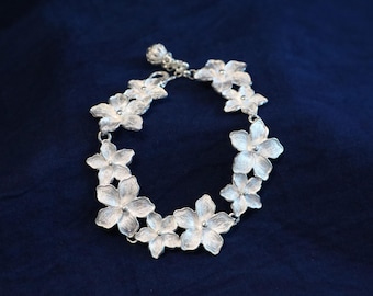 May include: A silver bracelet featuring a floral design. The bracelet is composed of multiple flower-shaped links, each with detailed petals and a central bead. The jewelry is displayed against a dark blue background.