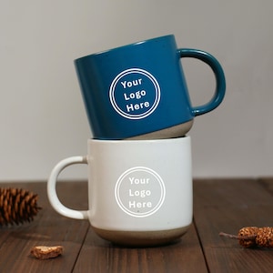 May include: Two ceramic mugs, one teal and one white, stacked on top of each other. Each mug has a rounded base and a curved handle. The teal mug has the text "Your Logo Here" in a white circle. The white mug also has the same text.