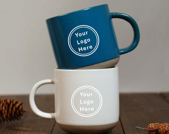 May include: Two ceramic mugs, one teal and one white, stacked on top of each other. Each mug has a rounded base and a curved handle. The teal mug has the text "Your Logo Here" in a white circle. The white mug also has the same text.