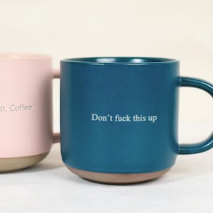 May include: Two ceramic mugs, one pink and one teal, with brown bases. The pink mug has the text "But First, Coffee" and the teal mug has the text "Don't fuck this up" in white. Both mugs have a rounded shape and a handle.