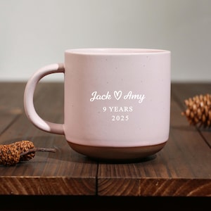 Może przedstawiać: Różowy ceramiczny kubek z brązową podstawą i uchwytem. Na kubku widnieje napis "Jack ♡ Amy" i "9 YEARS 2025". Kubek stoi na drewnianej powierzchni.