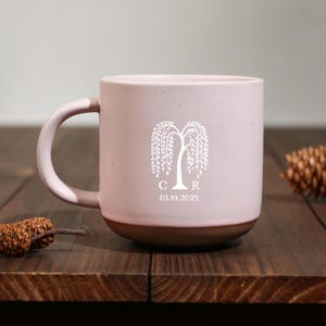 May include: A pink ceramic mug with a brown base and handle. The mug features a white weeping willow tree design with the initials "CR" and the date "03.14.2025". The mug is on a dark wooden surface.
