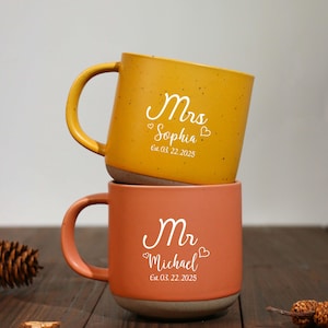 Pode incluir: Duas canecas de cerâmica empilhadas. A caneca superior é amarelo mostarda e diz "Mrs Sophia" com uma data. A caneca inferior é laranja e diz "Mr Michael" com uma data. Ambas as canecas têm uma textura salpicada.